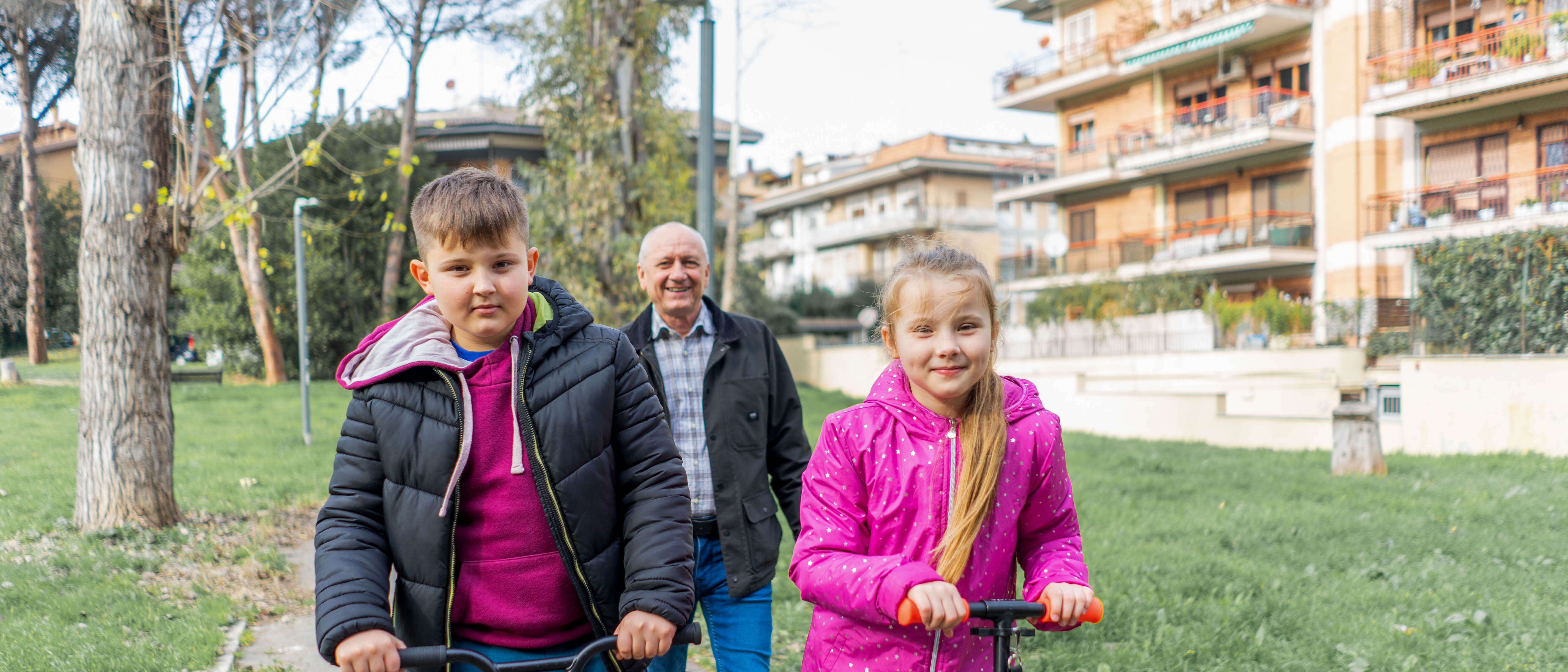 Two children ride scooters while their grandfather walks behind them.