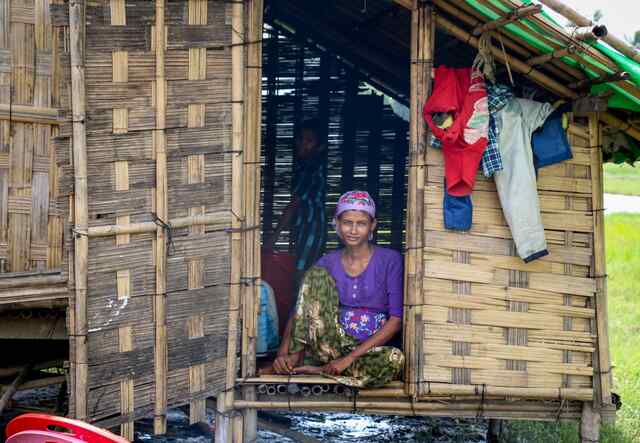 A woman sits in a small house on stilts, smiling softly towards the camera.