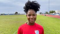 Wearing a red shirt and smiling, Kezia looks at the camera while on a soccer field 